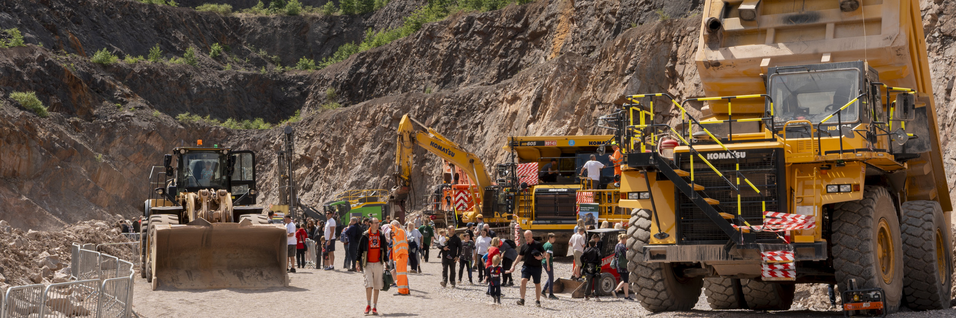 A quarry scene with large yellow construction vehicles, including a front loader on the left and a dump truck on the right. A group of people in safety gear stands in the center, suggesting a tour or inspection. The background features rocky, excavated walls with greenery at the top edges.