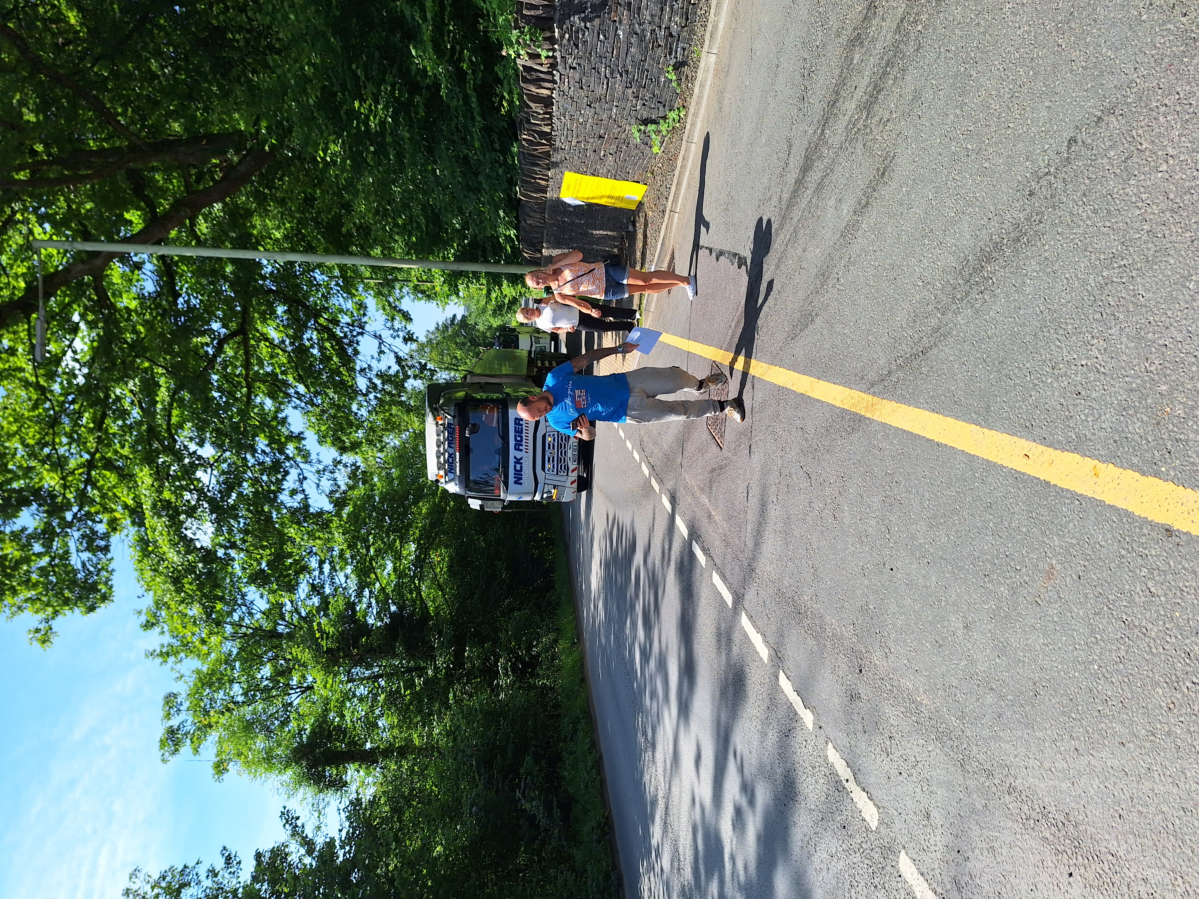 Two runners on a tree-lined road pass a stationary bus on the left side; one wears a blue shirt and the other a white tank top. A yellow centerline divides the asphalt road. Sunlight filters through the trees, creating dappled shadows.