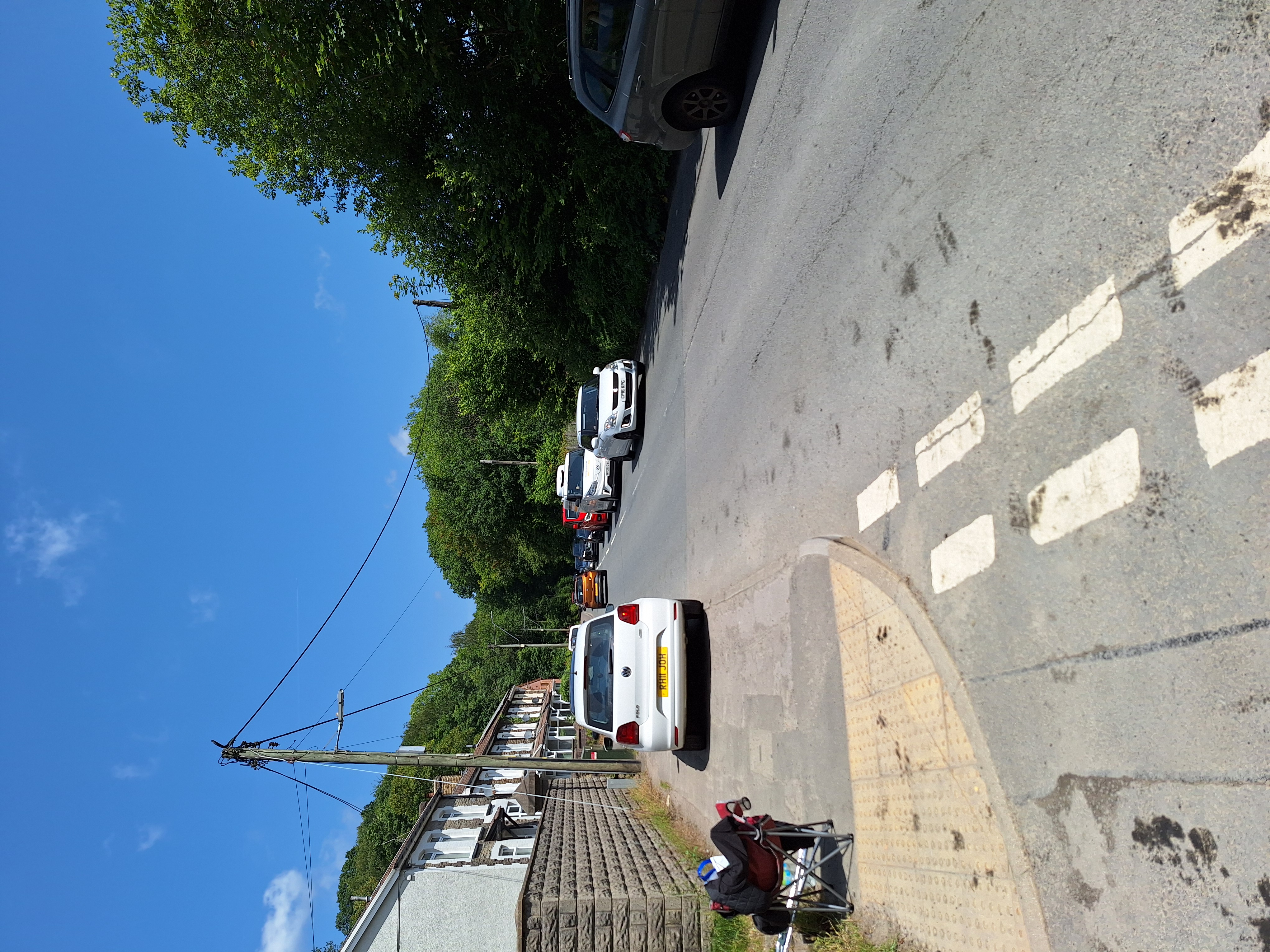 A pedestrian crossing at a quiet suburban road intersection on a sunny day. A white car is parked on the left side of the road near the crossing, while another vehicle approaches from the opposite side. Trees and houses line the left edge, with a mobility scooter visible on the pavement in the foreground.