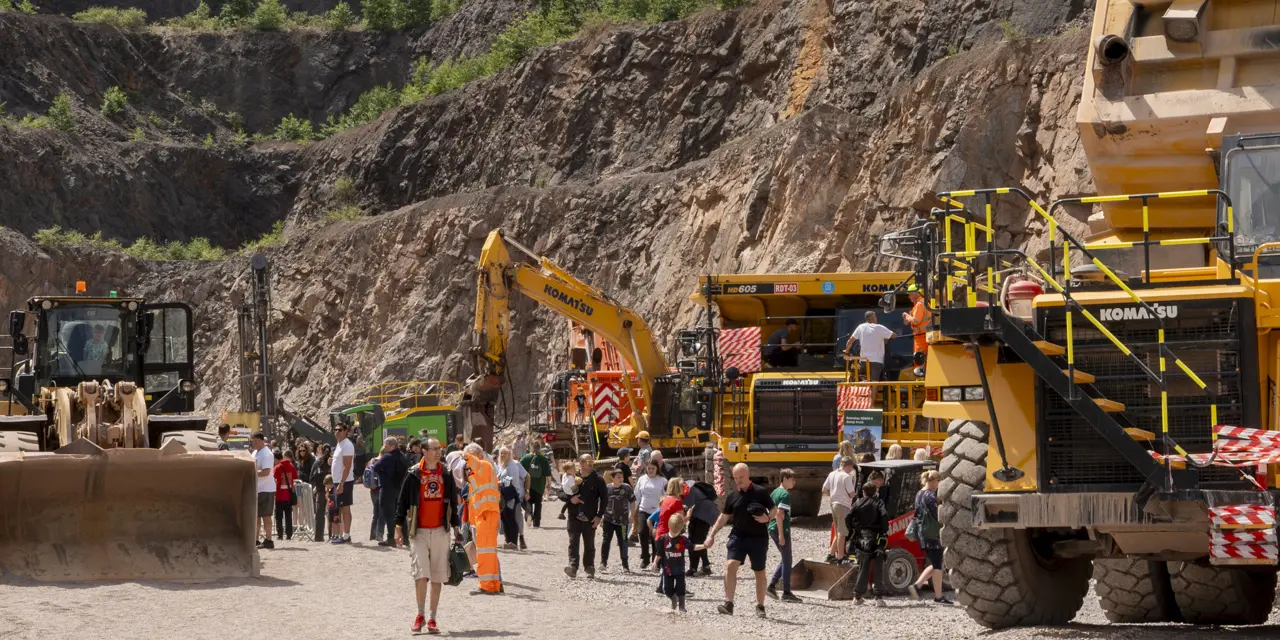 A quarry scene with large yellow construction vehicles, including a front loader on the left and a dump truck on the right. A group of people in safety gear stands in the center, suggesting a tour or inspection. The background features rocky, excavated walls with greenery at the top edges.