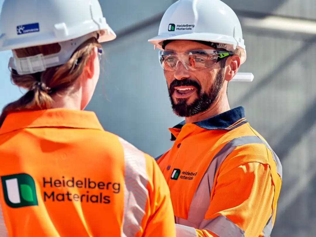 Heidelberg Materials workers wearing orange high-visibility jackets and white helmets in discussion at a worksite, with a logo reading "West Venture Holdings" on the back of one jacket; industrial equipment and a concrete structure visible in the background.
